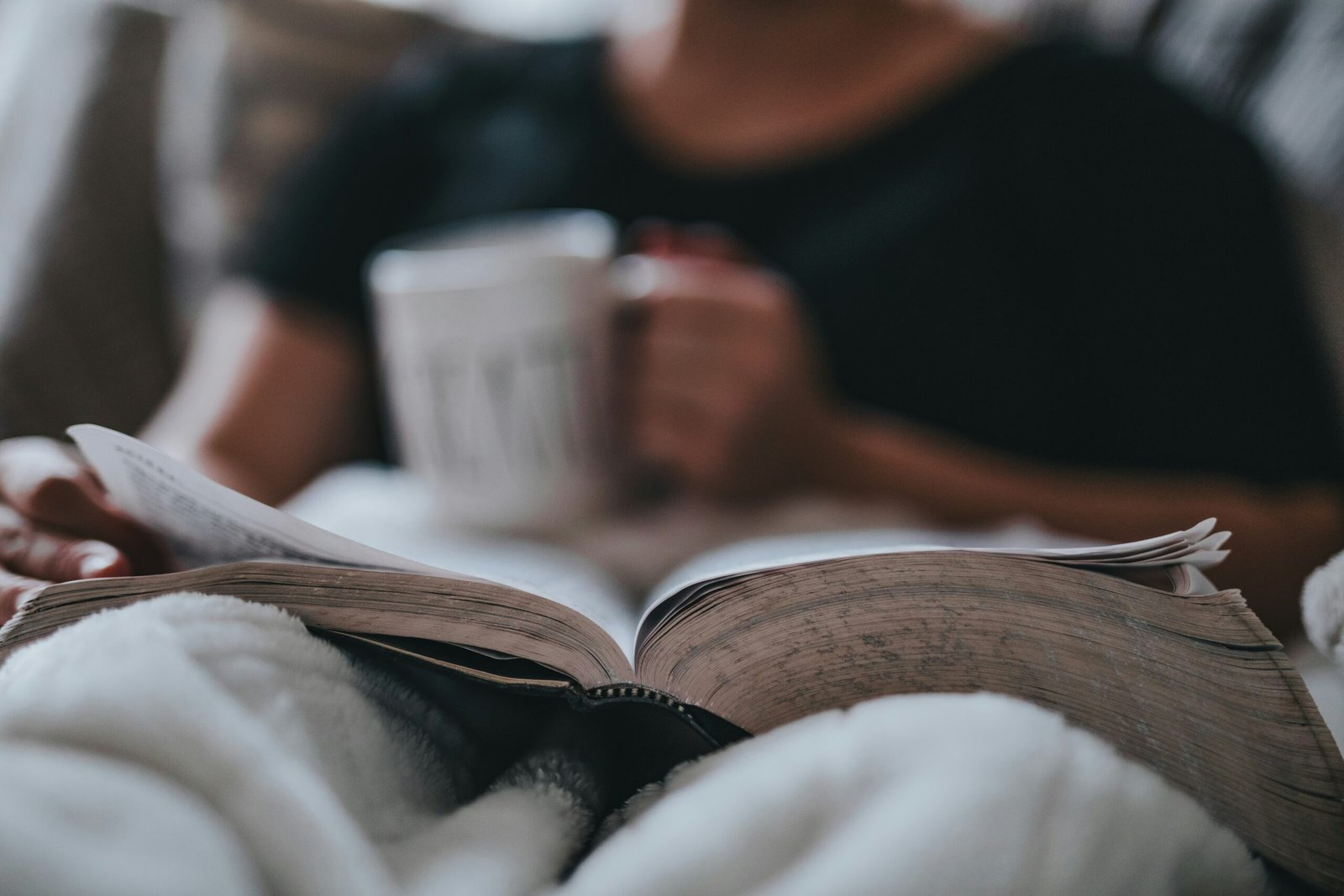 Person relaxing in bed with mug reading a book